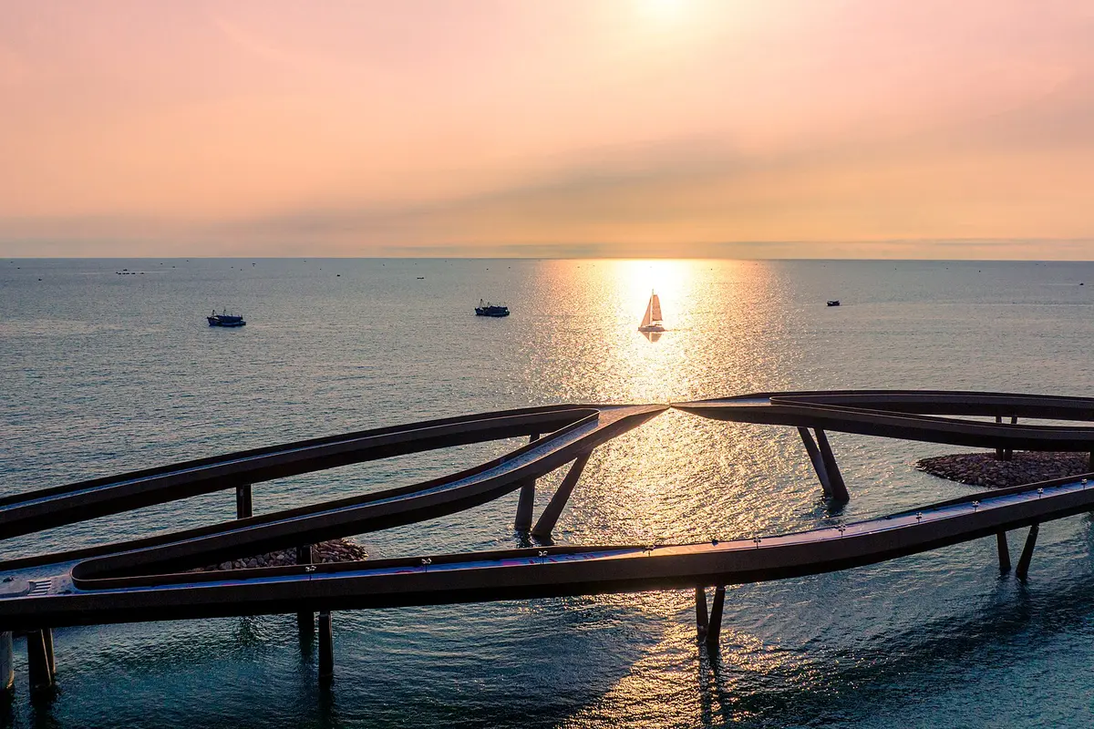 A curving walkway built out over the sea at sunset, with a sailing boat on the glittering water beyond